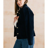 A woman wearing a dark navy corduroy jacket with gold buttons over a white ruffled shirt and blue jeans, standing against a light beige textured wall.