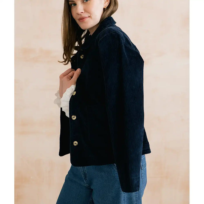 A woman wearing a dark navy corduroy jacket with gold buttons over a white ruffled shirt and blue jeans, standing against a light beige textured wall.