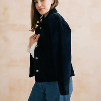 A woman wearing a dark navy corduroy jacket with gold buttons over a white ruffled long-sleeve shirt and blue jeans, standing against a warm beige textured wall.