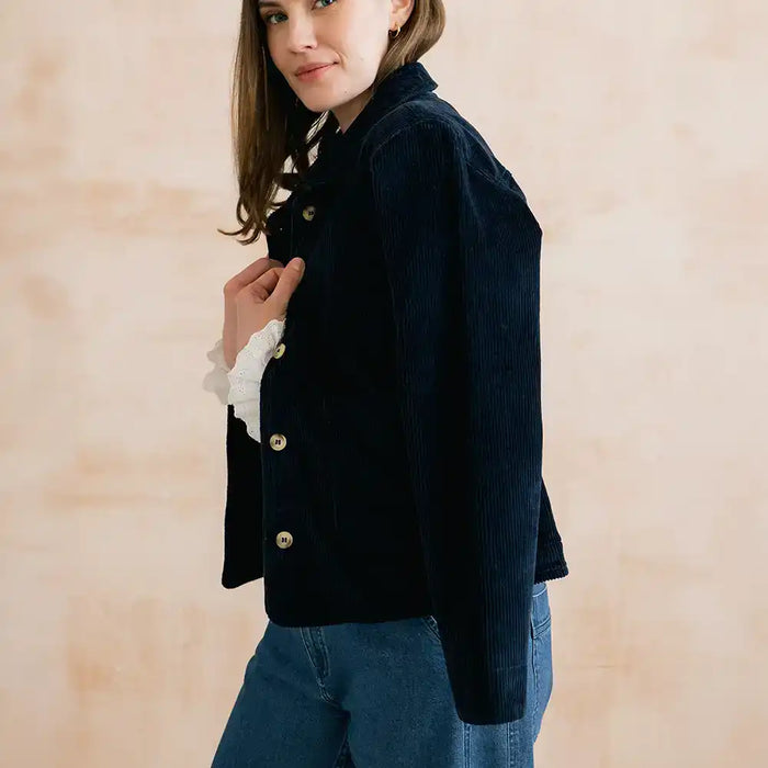 A woman wearing a dark navy corduroy jacket with gold buttons over a white ruffled long-sleeve shirt and blue jeans, standing against a warm beige textured wall.