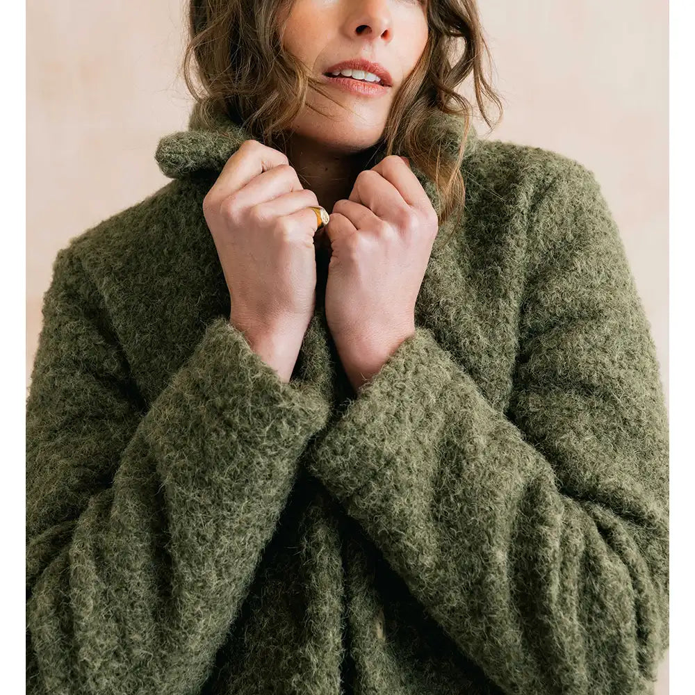 A close-up of a woman wearing a fuzzy, textured olive green knit sweater, holding the collar close with both hands against a soft beige background.