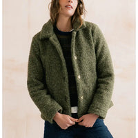 A woman wearing a fuzzy olive green cropped jacket over a dark striped sweater and blue jeans, standing against a light beige wall.