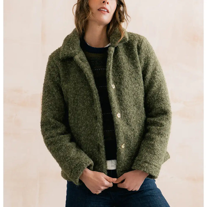 A woman wearing a fuzzy olive green cropped jacket over a dark striped sweater and blue jeans, standing against a light beige wall.