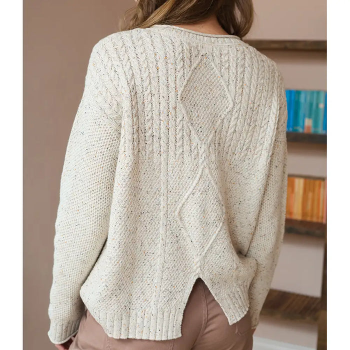 Beige cable knit sweater worn by a person, with a blurred indoor background.