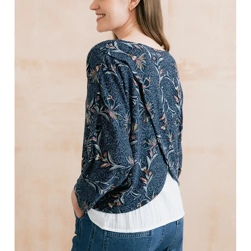 A woman wearing a dark blue patterned blouse with a white undershirt and blue jeans, shown from the back against a light beige background.