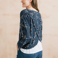 A woman shown from the back wearing a dark blue patterned blouse with a white undershirt and blue jeans, set against a soft beige background.