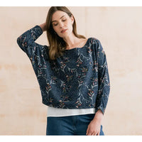 A woman wearing a navy blue long-sleeve blouse with a delicate floral and botanical print, styled with a white ribbed undershirt and blue jeans, set against a warm beige textured wall.