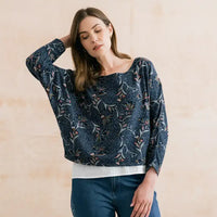 A woman wearing a navy blue long-sleeve blouse with a white and pink floral pattern, paired with blue jeans, posing against a neutral beige wall.