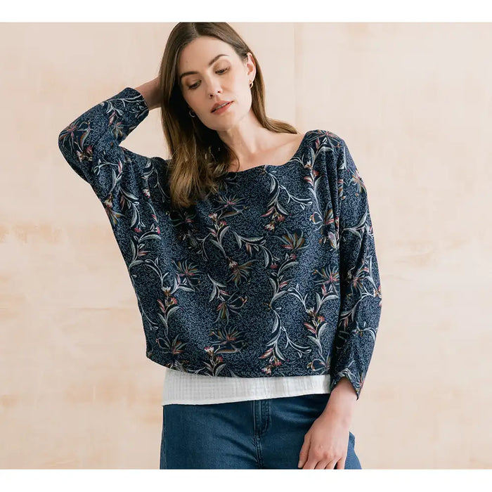A woman wearing a navy blue long-sleeve blouse with a delicate floral and botanical print, styled with a white ribbed undershirt and blue jeans, set against a warm beige textured wall.
