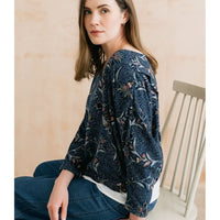 A woman wearing a dark navy blue long-sleeve blouse with a dense floral and botanical print in muted orange, beige, and green tones, paired with blue jeans, seated on a light-colored wooden chair.