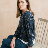 A woman wearing a dark navy blue long-sleeve blouse with a dense floral and botanical print in muted green, pink, and beige tones, paired with blue jeans, seated on a wooden chair against a warm beige wall.