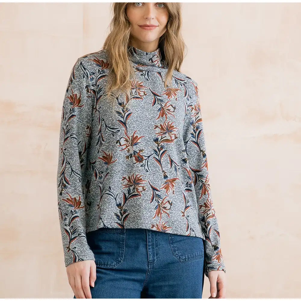 A woman wearing a gray mock-neck long-sleeve top with a brown and orange floral pattern, paired with blue jeans, standing against a light beige textured wall.