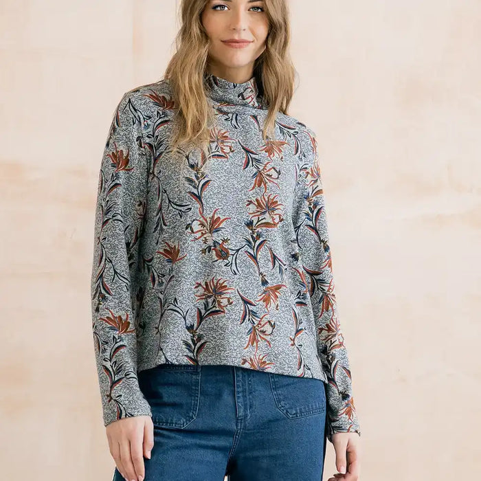 A woman wearing a gray mock-neck long-sleeve top with a rust-orange and brown floral pattern, paired with blue jeans, standing against a light beige background.
