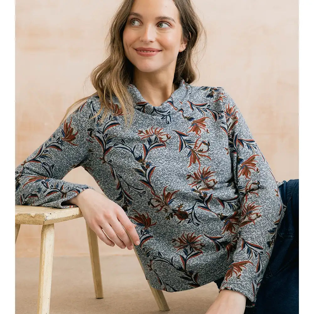 A woman wearing a long-sleeve ruffled blouse with a gray and white floral pattern and rust-orange accents, leaning on a wooden stool against a warm beige background.
