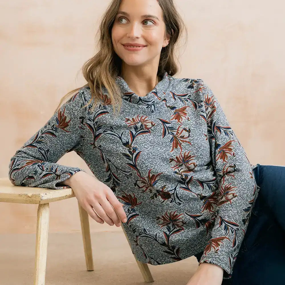 A woman wearing a long-sleeve button-up blouse with a gray base and a brown and black floral pattern, leaning on a wooden stool against a warm beige background.