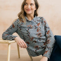 A woman wearing a long-sleeve button-up blouse with a gray base and a brown and black floral pattern, leaning on a wooden stool against a warm beige background.