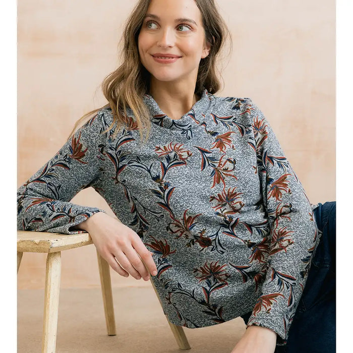 A woman wearing a long-sleeve ruffled blouse with a gray and white floral pattern and rust-orange accents, leaning on a wooden stool against a warm beige background.