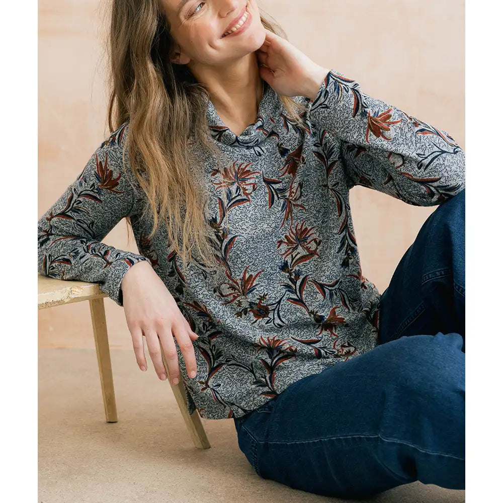 A woman wearing a long-sleeve gray patterned blouse with brown and black botanical motifs, paired with dark blue jeans, sitting on a beige floor next to a wooden stool.