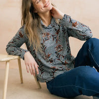 A woman wearing a long-sleeve patterned shirt and blue jeans sits on a beige floor next to a light wood stool, looking up and smiling.