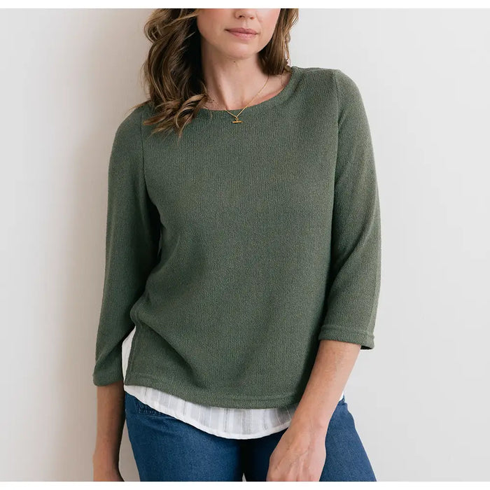 Woman wearing a green sweater over a white shirt with blue jeans against a plain background