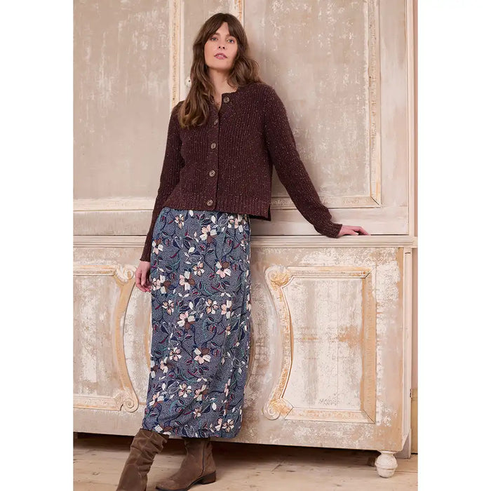 Woman wearing a brown cardigan and floral skirt standing against an ornate wall.