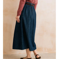 A person wearing a dark blue denim midi skirt with a brown belt, paired with a dusty rose long-sleeve top and brown flat sandals, standing against a textured beige wall.