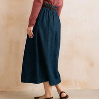 A person wearing a dark blue denim midi skirt with a brown belt, paired with a dusty rose long-sleeve top and dark brown flat sandals, standing against a textured beige wall.