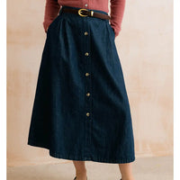 A dark blue denim midi skirt with a button-down front, styled with a brown leather belt and gold buckle, worn by a model in a dusty rose top against a beige wall.