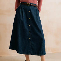 A dark blue denim midi skirt with a button-down front, styled with a brown belt and a rust-colored top, shown on a model against a textured beige wall.