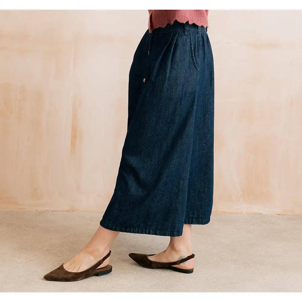 A person wearing dark blue denim culotte-style pants with a scalloped waistband and front button detail, paired with dark brown slingback flats, standing against a warm beige wall.