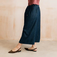 A person wearing dark blue denim culottes with a scalloped hem and front button detail, paired with a pink top and dark brown slingback flats, standing against a warm beige wall.