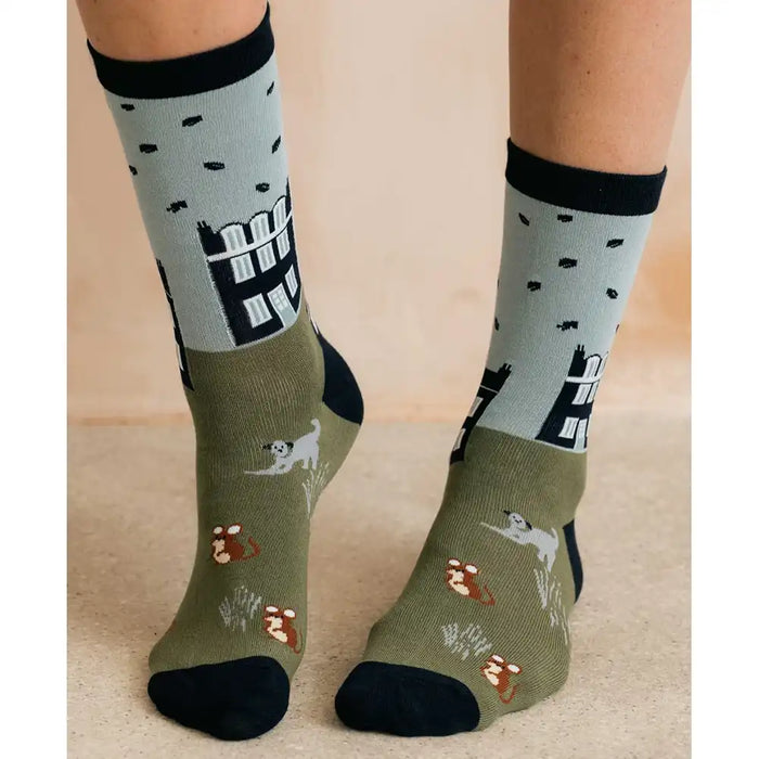 A pair of crew socks worn on a person's feet, featuring an olive green base with black and white architectural illustrations, small white dogs, and brown-and-white foxes, set against a warm beige background.