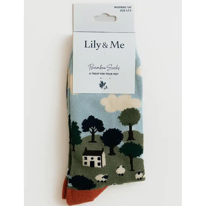 A pair of packaged Bamboo Socks by Lily & Me, featuring a printed landscape design with trees, sheep, a small house, and clouds on a light blue background, with rust-orange toes.