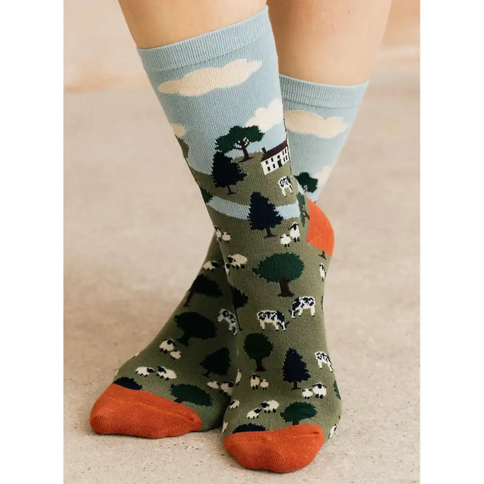 A pair of crew socks worn on feet, featuring a farm landscape pattern with cows, trees, and a barn on an olive green background, with a light blue upper section and orange toes and heels.
