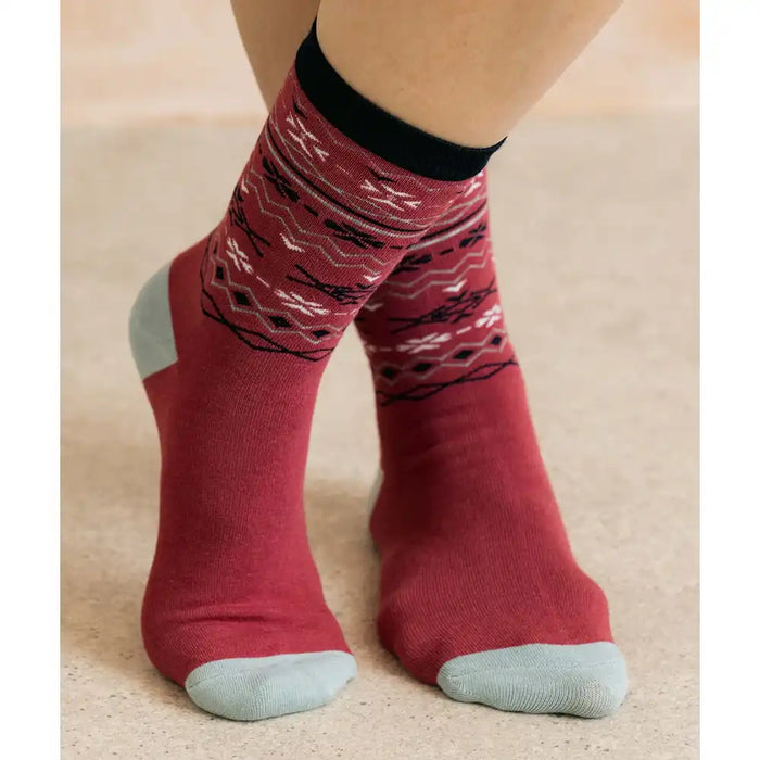 A pair of crew socks worn on feet, featuring a rust red body with a black, white, and gray geometric pattern on the upper half, a solid black cuff, and light gray toes and heels, shown against a neutral beige background.