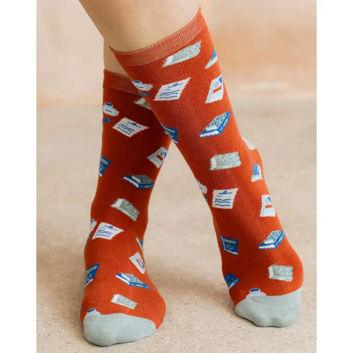 A pair of crew socks worn on a person's feet, featuring an orange base with a pattern of blue and white books and pages, set against a neutral beige background.