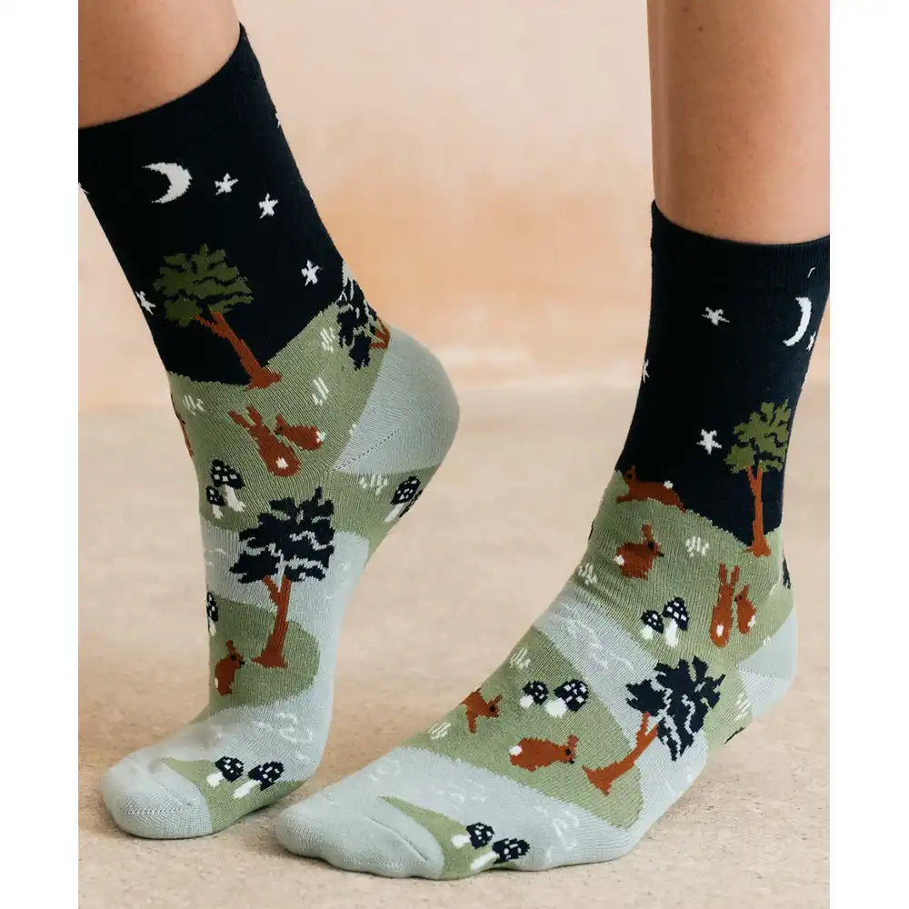 A pair of crew socks worn on feet, featuring a woodland forest pattern with trees, rabbits, mushrooms, and a night sky design in black at the top.