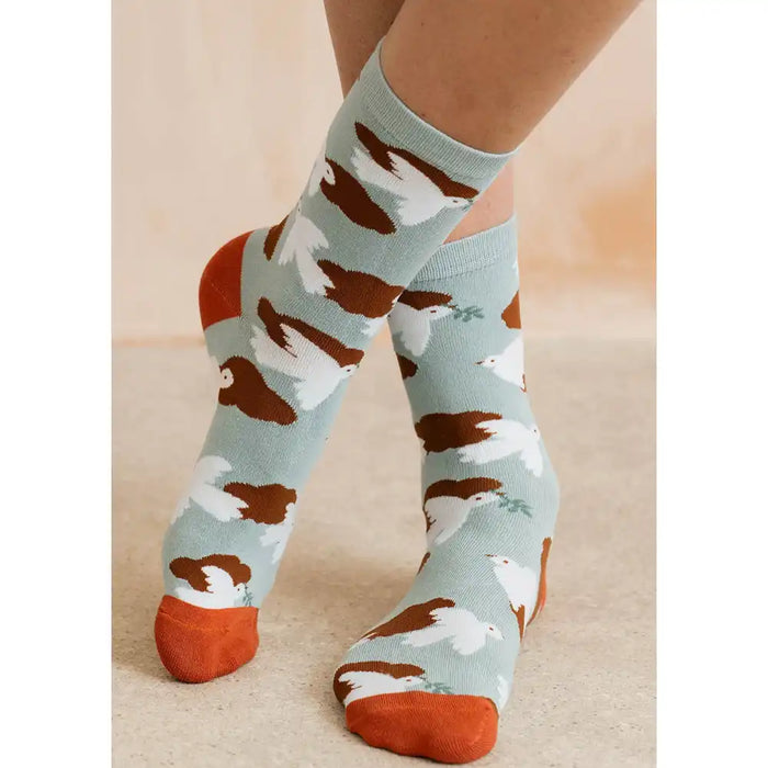 A person wearing crew socks with a pattern of brown and white birds on a pale blue background, with orange heels, toes, and cuffs, shown against a neutral beige floor and wall.