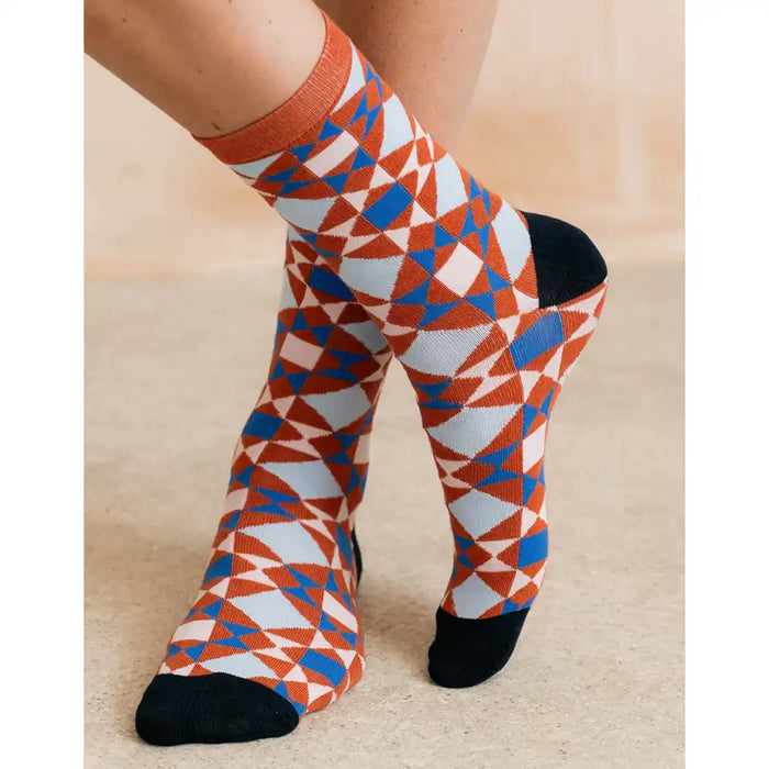 A pair of crew socks with a geometric pattern in orange, blue, white, and black, worn on a person's legs against a neutral beige background.
