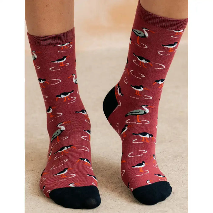 A pair of crew socks worn on a person's feet, featuring a pattern of black-and-white birds with orange legs on a dusty rose background, with solid black toes and heels.