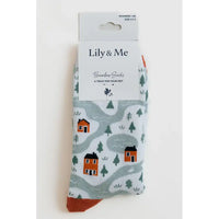 A pair of Lily & Me bamboo socks in a white paper packaging sleeve, featuring a winter landscape pattern with orange houses, green trees, and gray winding paths on a white background.