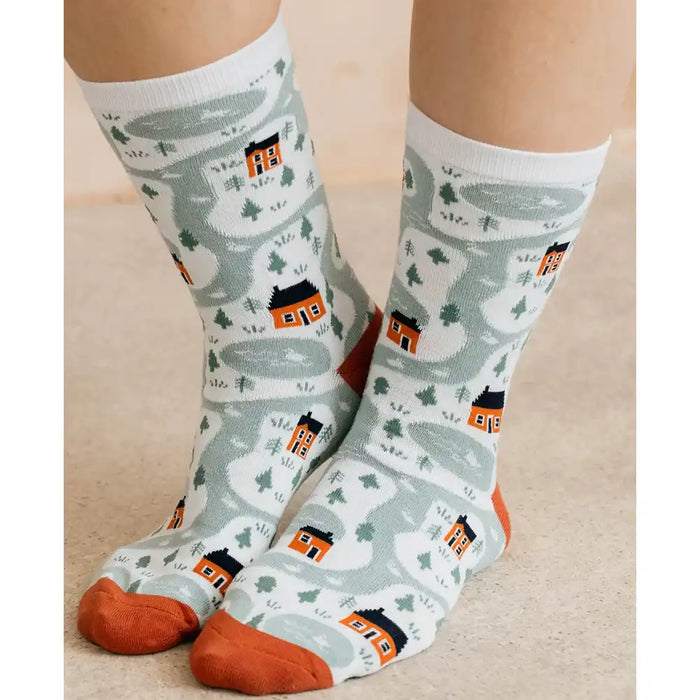 A pair of crew socks worn on a person's legs, featuring a whimsical winter landscape pattern with winding gray paths, small orange houses, and pine trees on a white background, with orange toes and heels.
