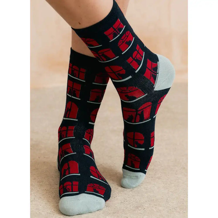 A person's feet wearing black crew socks with a red and gray geometric pattern, standing on a beige floor against a neutral beige wall.