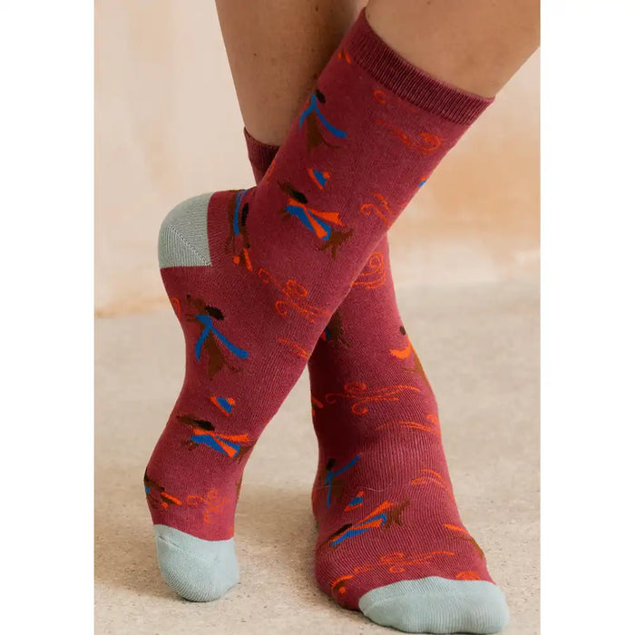 A close-up of a person's feet wearing crew socks with a burgundy base and a pattern of small blue, orange, and brown birds, finished with light gray heels, toes, and cuffs.