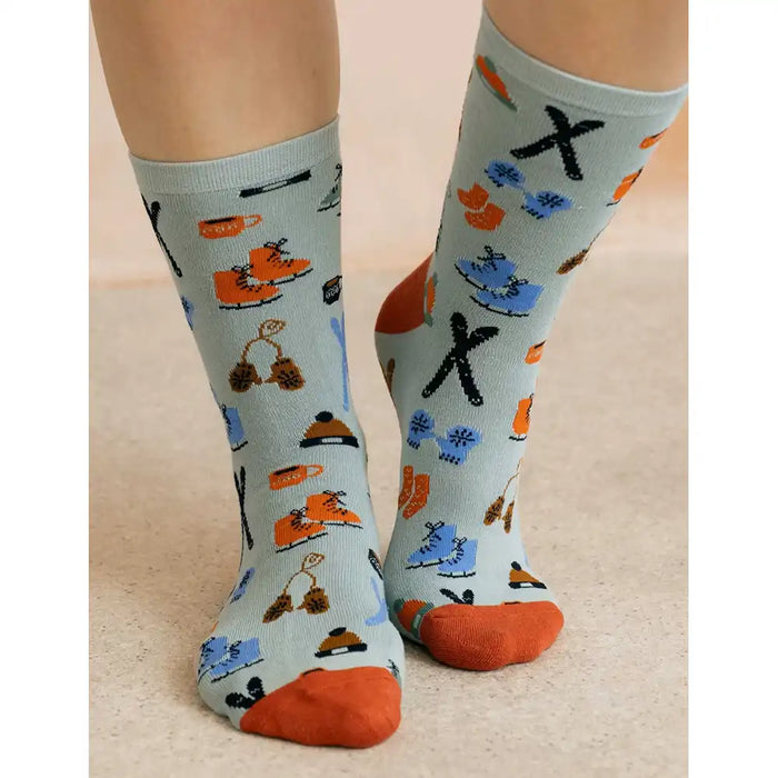 A pair of crew socks worn on feet, featuring a light blue-gray background with a playful pattern of orange skates, blue snowboards, black and orange winter hats, and orange toes and heels.
