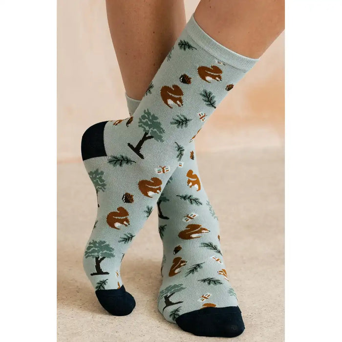 A pair of crew socks with a woodland forest pattern featuring brown squirrels, pine trees, and acorns on a light sage green background, with solid black heels, toes, and cuffs, shown on a model's feet.