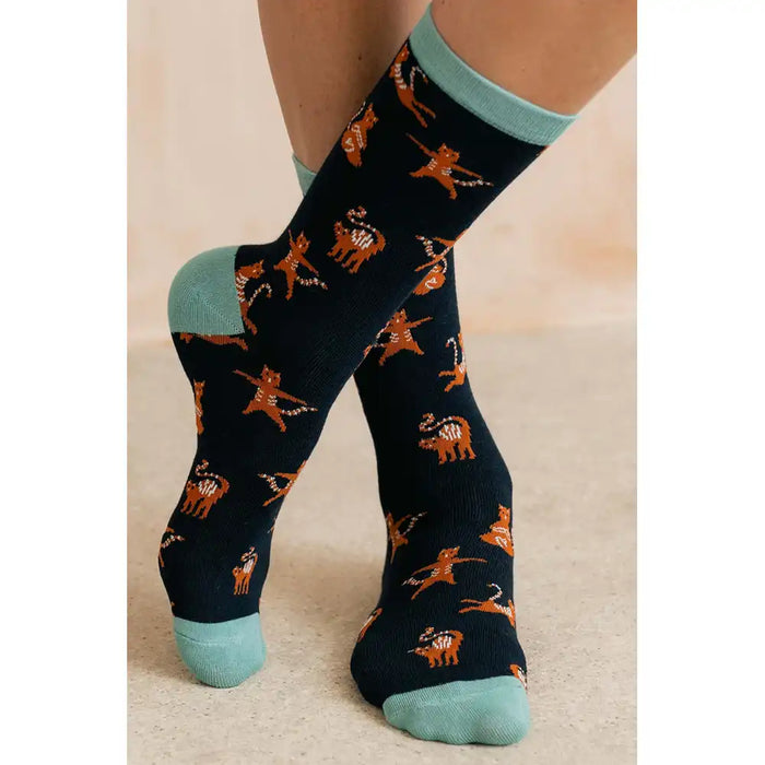A pair of crew socks worn on a person's feet, featuring a dark navy blue base with an all-over pattern of orange and white dancing cats, accented with light mint green cuffs, heels, and toes.