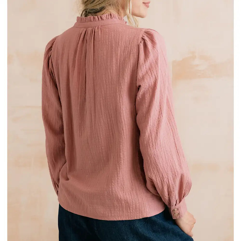 A woman wearing a dusty rose crinkle-textured blouse with puff sleeves and a ruffled high neckline, shown from the back against a warm beige background.