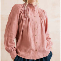 A woman wearing a dusty rose pink long-sleeve blouse with a high ruffled collar, vertical pleats on the chest, and a button-down front, paired with dark blue jeans against a warm beige background.