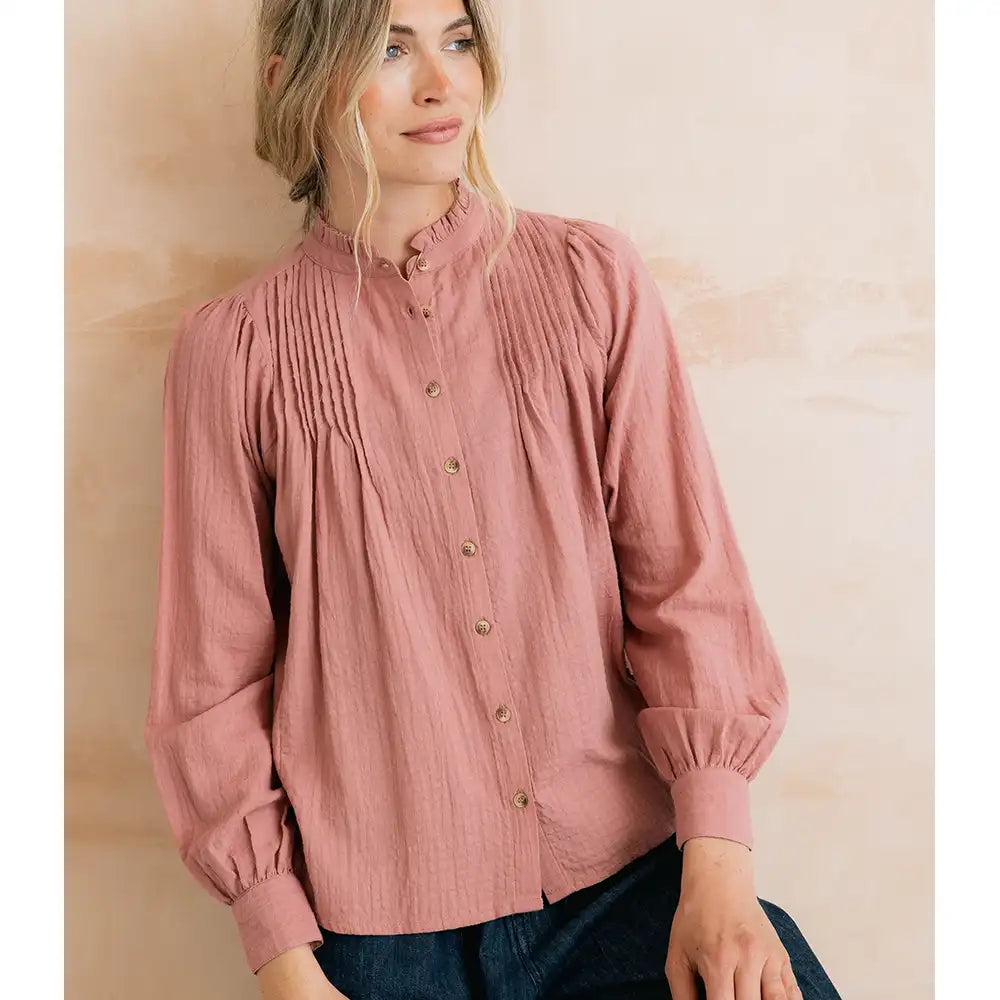 A woman wearing a dusty rose long-sleeve blouse with a high ruffled collar, gathered front, and button closure, paired with dark jeans against a soft beige textured wall.